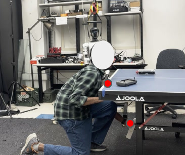 A person kneels at a table-tennis table, one hand on the table edge and one reaching underneath; red dots mark the contacts. Face blurred.
